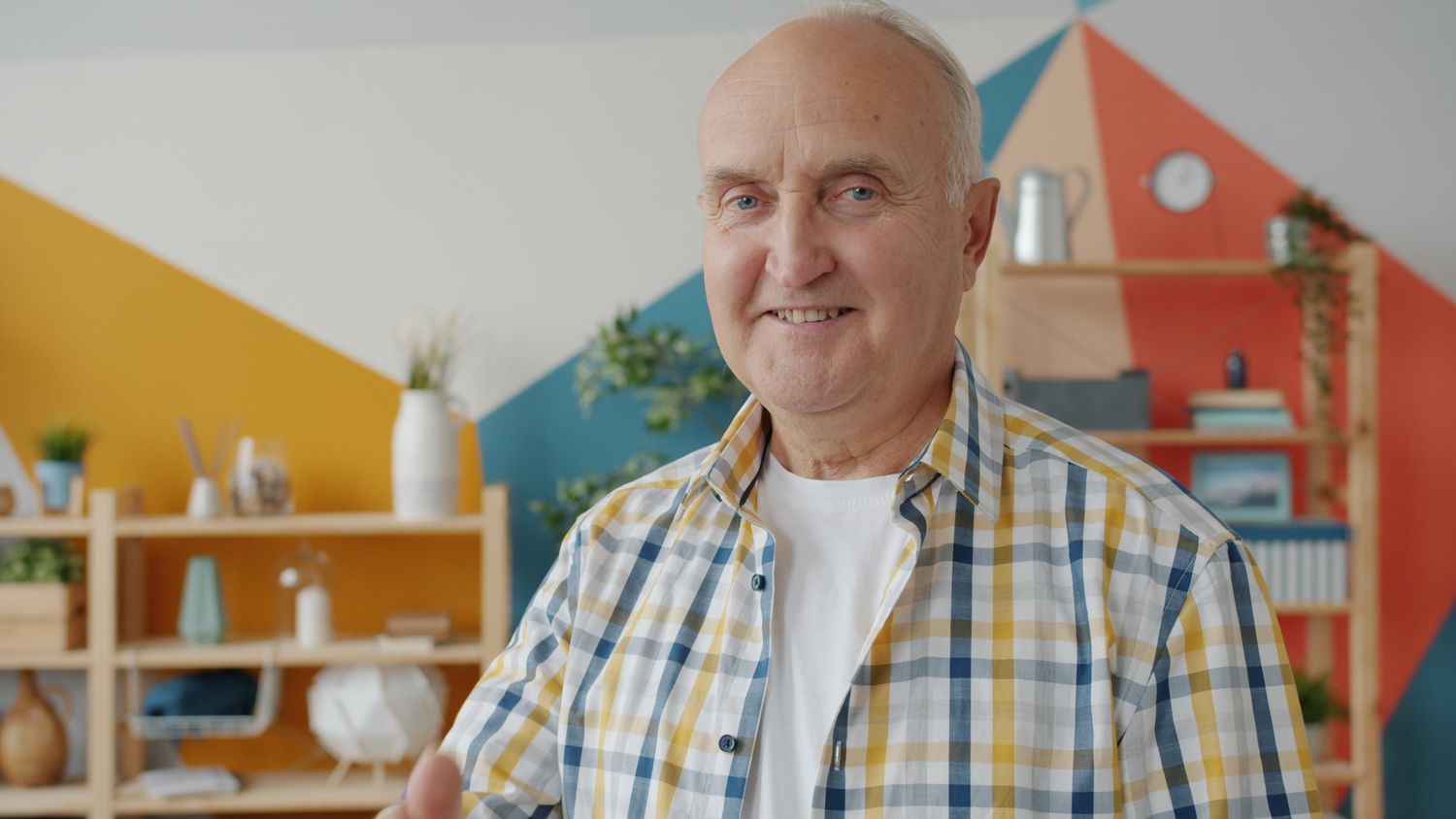An older man in a plaid shirt smiles at the camera in a bright room at home.