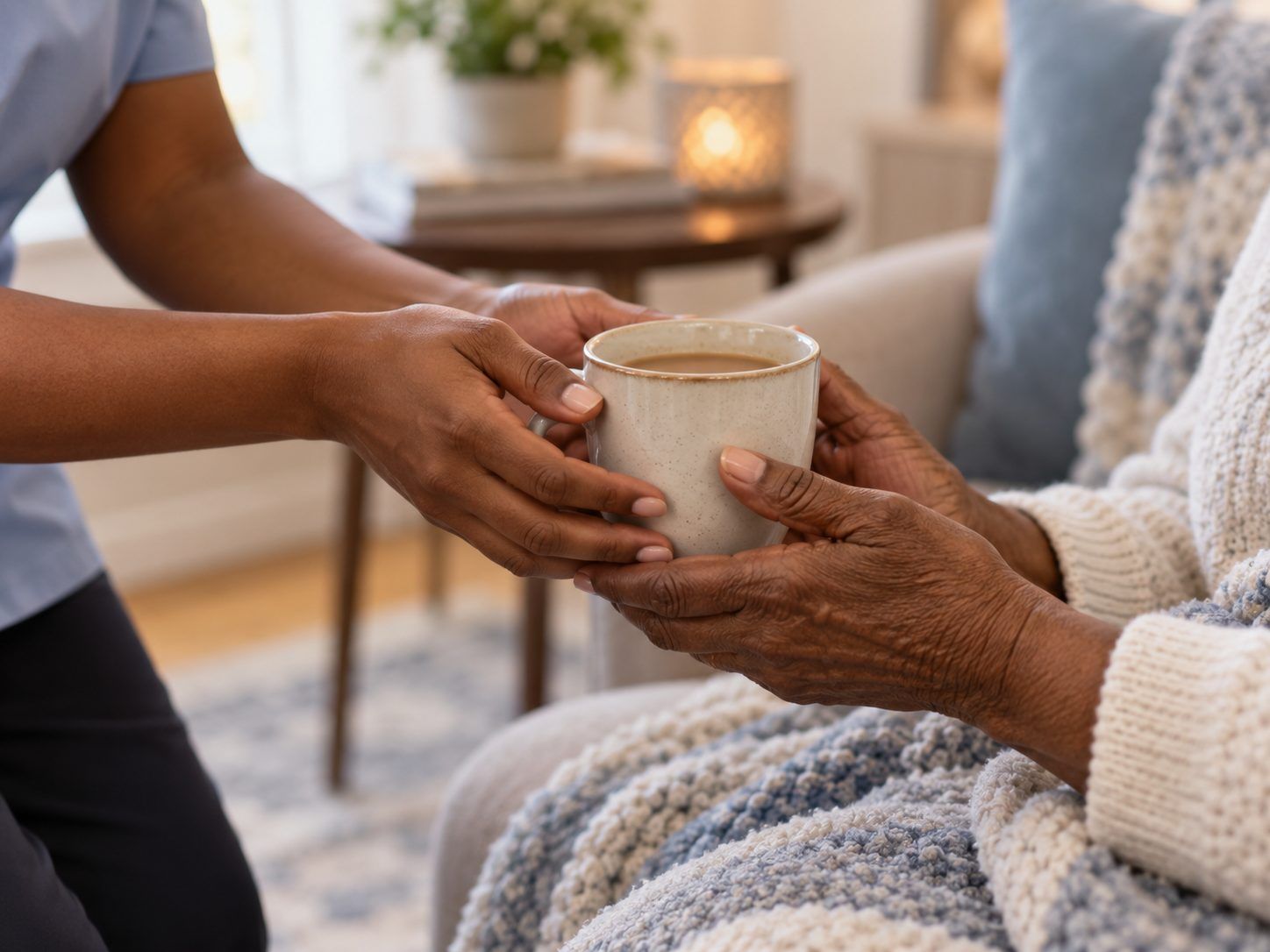 A caregiver's hands pass a warm mug to an older person wrapped in a knit blanket.
